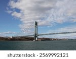 A suspension bridge extending over a wide body of water, with an urban shoreline and overcast sky in the background, highlighting the bridge