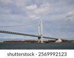 A suspension bridge extending over a wide body of water, with an urban shoreline and overcast sky in the background, highlighting the bridge