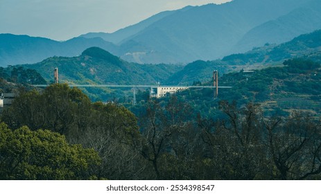 A suspension bridge spans across lush, forested hills with a misty backdrop of towering mountains. - Powered by Shutterstock