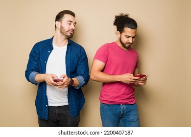 Suspicious Jealous Gay Man Spying On His Boyfriend Texting On His Smartphone In Front Of A Yellow Background