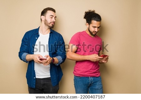 Suspicious jealous gay man spying on his boyfriend texting on his smartphone in front of a yellow background