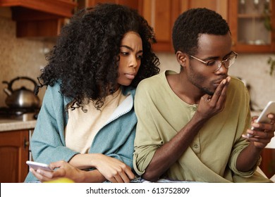 Suspicious Jealous Young African American Wife Looking Over Her Husband's Shoulder, Reading Messages On Mobile Phone While He Is Texting Someone, Covering Mouth With Hand, Having Worried Look