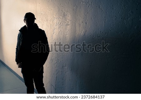 Suspicious man. Silhouette of criminal. Stalker or anonymous stranger. Gang crime, teenage gangster or thief. Dark urban street alley. Face hidden in shadow. Scary figure at night. Grunge background.