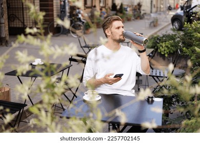 Sustainability city. Man drinks tap water from reusable metal bottle. Green city street cafe. Man wearing white shirt and using smartphone. Coffee cup on the table. Workation mobile internet on the go - Powered by Shutterstock