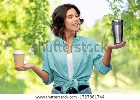 sustainability, eco and green living concept - portrait of happy smiling young woman in turquoise shirt comparing thermo cup or tumbler with disposable paper coffee cup over natural background
