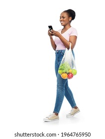 Sustainability, Food Shopping And Eco Friendly Concept - Happy Smiling African American Woman With Smartphone Holding Reusable String Bag With Fruits And Vegetables Walking Over White Background