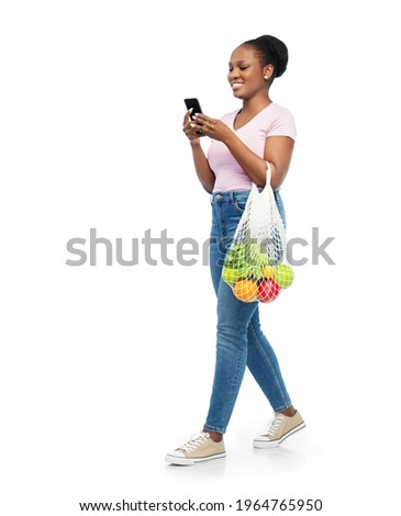 sustainability, food shopping and eco friendly concept - happy smiling african american woman with smartphone holding reusable string bag with fruits and vegetables walking over white background