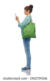 Sustainability And People Concept - Happy Woman In Turquoise Shirt And Jeans With Smartphone And Green Reusable Canvas Bag For Food Shopping Over White Background