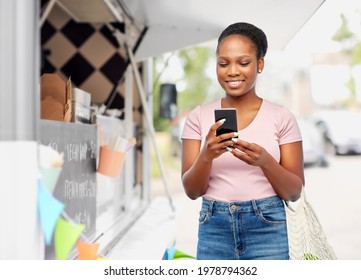 Sustainability, Shopping And Eco Friendly Concept - Happy Smiling African American Woman With Smartphone Holding Reusable String Bag Over Food Truck Background