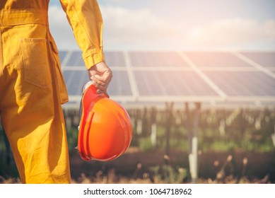 Sustainable And Clean Energy Concept. Construction Engineer Or Electrical Worker Holding Orange Safety Helmet Work At Solar Panels Background. Foreman Wearing Safety Suit And Looking At Power Plant