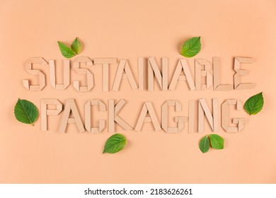 Sustainable Paper Packaging Concept. Text Sustainable Packaging With Green Leaves Around Over Light Brown Background. Flat Lay Style