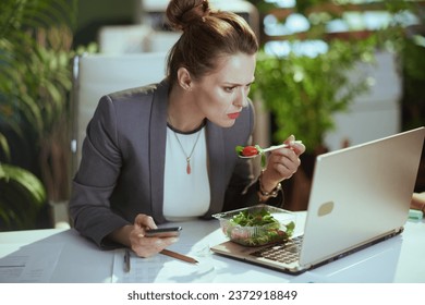 Sustainable workplace. concerned modern middle aged bookkeeper woman in a grey business suit in modern green office with laptop eating salad and using smartphone.