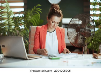 Sustainable workplace. modern 40 years old bookkeeper woman in modern green office in red jacket with electric fan and laptop. - Powered by Shutterstock