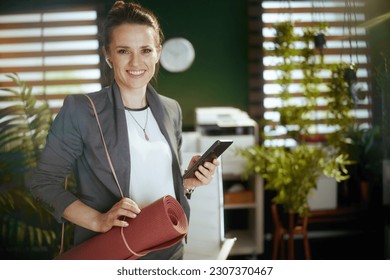 Sustainable workplace. smiling modern small business owner woman in modern green office with wireless headphones, yoga mat and smartphone. - Powered by Shutterstock