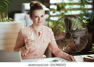 Sustainable workplace. smiling modern woman worker in modern green office with electric fan, cup of water and lemon.