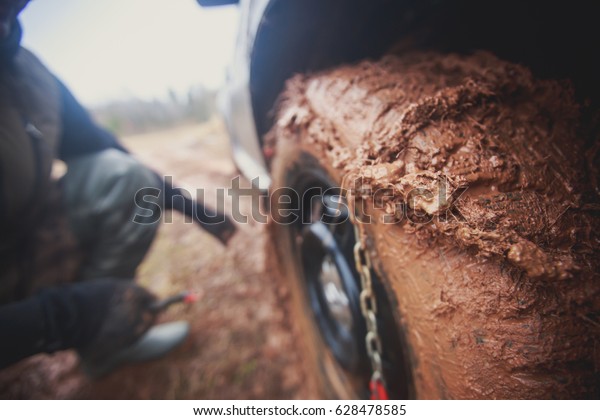 Suv 4wd car stuck\
in muddy off-road track road field with a wheel covered in mud\
terrain and a man trying to put chains on it in the background\
during jeeping competition