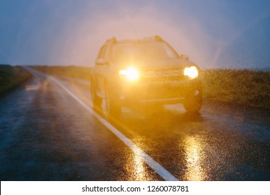 SUV Car On Wet Asphalt Road Drives In Dark Rainy Conditions. Out Of Focus Countryside Landscape With Raindrops In The Rays Of Headlights