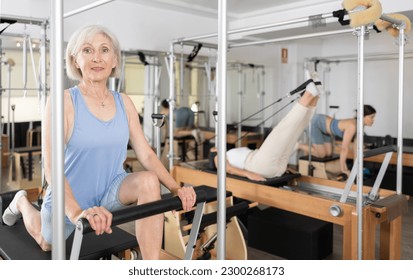 Svelte elderly woman performing set of pilates exercises on reformer during group workout. Active lifestyle and wellness concept of seniors.. - Powered by Shutterstock