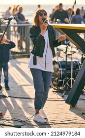 Svetlogorsk, Russia - 08.15.2021 - Young Woman In Black Leather Jacket Singing Song In Microphone At Outdoor Rock Festival, Sea Background. Rock Singer Performing At Open Air Concert, Sunny Day