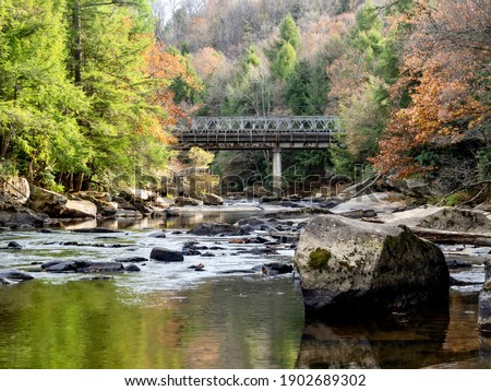 Swallow Falls State Park in Maryland with the water in the foreground, large rocks, and fall foliage trees and a bridge in the background, with colors reflecting in the water.  Beautiful nature.