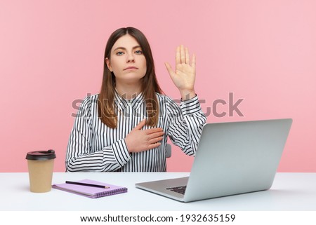 I swear! Honest dedicated woman office worker raising hand to give promise sitting at workplace, taking oath with trustworthy faithful expression. Indoor studio shot isolated on pink background