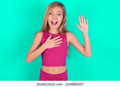 I Swear, Promise You Not Regret. Portrait Of Sincere Blonde Little Kid Girl Wearing Pink Sport Clothes Over Green Background Raising One Arm And Hold Hand On Heart As Give Oath, Telling Truth, 