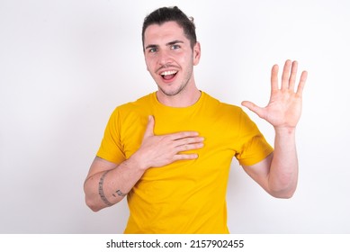 I Swear, Promise You Not Regret. Portrait Of Sincere Young Caucasian Man Wearing Yellow T-shirt Over White Background Raising One Arm And Hold Hand On Heart As Give Oath, Telling Truth,