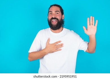 I Swear, Promise You Not Regret. Portrait Of Sincere Caucasian Man With Beard Wearing White T-shirt Over Blue Background Raising One Arm And Hold Hand On Heart As Give Oath, Telling Truth, Want You 