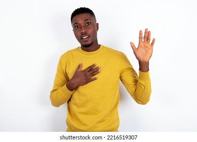 I Swear, Promise You Not Regret. Portrait Of Sincere Young Handsome Man Wearing Yellow Sweater Over White  Background Raising One Arm And Hold Hand On Heart As Give Oath, Telling Truth, Want You To Be