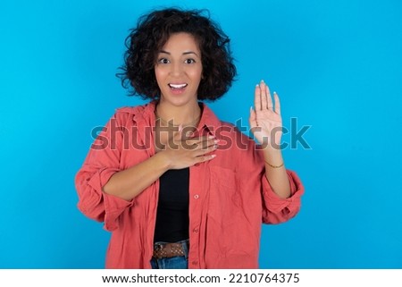 I swear, promise you not regret. Portrait of sincere young beautiful woman with curly short hair wearing red overshirt over blue wall raising one arm and hold hand on heart as give oath, telling truth