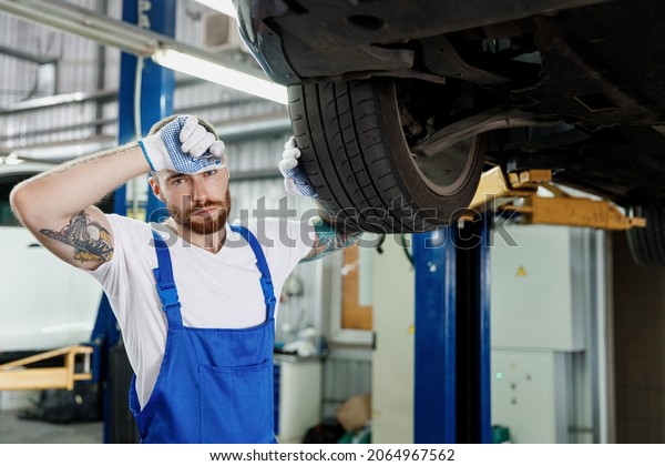 Sweating repairman tired young male professional\
technician mechanic man wears denim blue overalls white t-shirt\
stand near car lift changes wheel tires work in vehicle repair shop\
workshop indoors