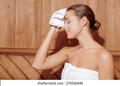 Sweating In Sauna. Young Woman Feels The Heat In Sauna Room And Cleans Sweat From Her Face With Small White Towel 