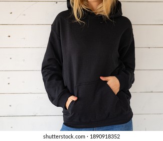 Sweatshirt hoodie mockup. Unrecognizable woman poses in a black sweatshirt against the background of white boards, facing the camera.