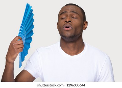 Sweaty African Man Suffering From Heat Stroke Problem Holding Waving Using Fan Isolated On White Grey Studio Background, Funny Black Guy Sweating Cooling Himself Feeling Relief In Summer Hot Weather