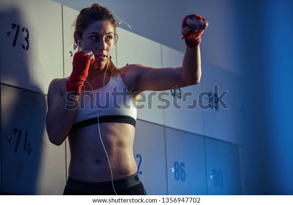 Sweaty athletic woman in fighting stance preparing for\
boxing training while listening music on earphone in gym\'s dressing\
room. 