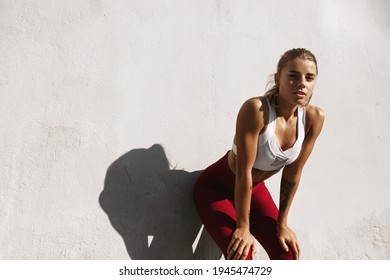 Sweaty fitness girl taking breath on morning jogging. Female athlete runner resting, workout outdoors on fresh air, jogger take break, wearing sports outfit, standing near concrete wall - Shutterstock ID 1945474729