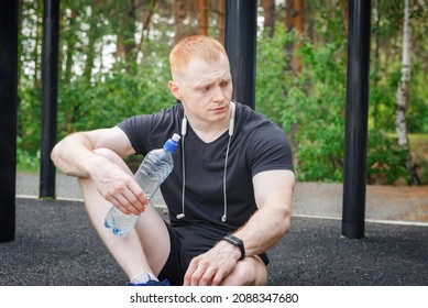 Sweaty Guy Opening Cap Of Bottle To Drink Water After Workout. Tired Man Resting After Training And Holding Bottle Of Water. Outdoor Training In The Park.