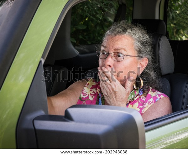 Sweaty, hot, senior woman driver looking\
terrified holding hand to mouth in driver\'s\
seat.