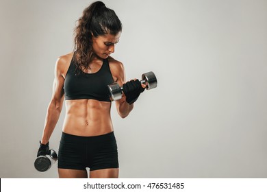 Sweaty muscular woman with pony tail hair style and shorts curling dumbbell over gray background with copy space