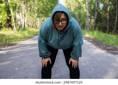 A sweaty woman looks at the camera with her hands on her knees and catching her breath after a run. An overweight woman breathes heavily after running. Break and rest after running. - Powered by Shutterstock