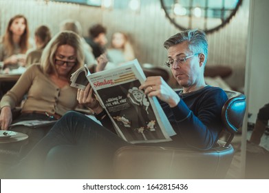 Malmö / Sweden - 09 February 2019:  A Casual Clothed Swedish Man In His 40s Enjoying The Weekend Reading A Sunday Newspaper In A Cozy Espresso House Cafe In The City Centre Of Malmo With His Partner.