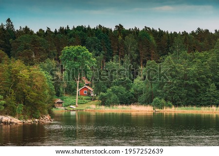 Sweden. Beautiful Red Swedish Wooden Log Cabin House On Rocky Island Coast In Summer. Lake Or River And Forest Landscape.