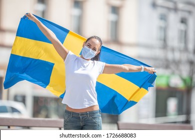 Swedish female fan in a facemask holds a flag behind her back.