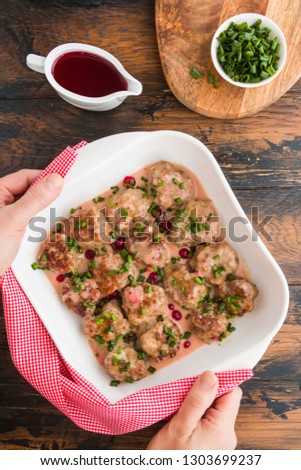 Swedish meatballs in a red cowberry sauce with green onion. White casserole on wooden rustic table, woman hands, top view 