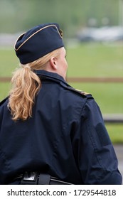 A Swedish Police (female) With Her Back Turned And Blonde Hair In A Pony Tail In Front Of A Blurred Bakground.