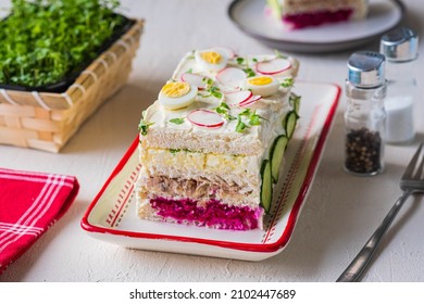 Swedish sandwich cake stuffed with egg salad, liver pate and beetroot salad, decorated with boiled quail eggs, slices of radish, cucumber and microgreens, on a green background.