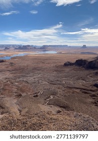 A sweeping elevated view across an expansive red desert plateau with winding blue waterways, distant flat-topped mesas, and dramatic rock formations stretching to the horizon under a partly cloudy sky.
