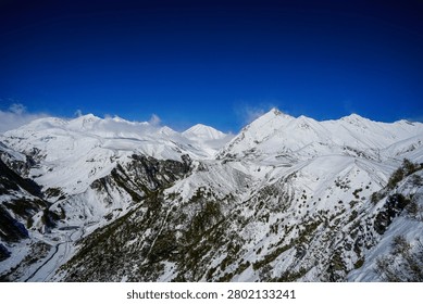 A sweeping panoramic view captures the vast, snow-covered expanse of the Greater Caucasus Mountains and the active ski resort slopes near Gudauri, Georgia. Deep valleys with evergreen trees and winding paths cut through the pristine white landscape under a brilliant, cloudless blue sky.