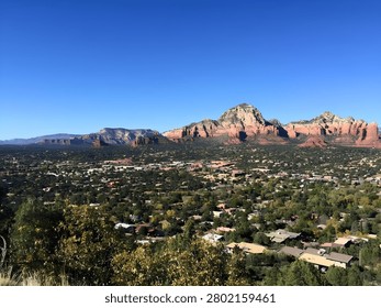 A sweeping panoramic view of a desert mountain landscape, where a quiet town nestles among dense green vegetation beneath towering red sandstone cliffs. The layered mesas and rugged peaks glow in warm sunlight against a vast, cloudless blue sky, creating a striking contrast between the arid rock formations and the lush valley below. The scene captures the timeless beauty of the American Southwest, with dramatic geology, open space, and a peaceful residential landscape.