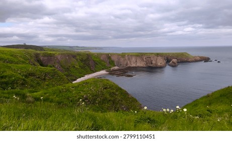 A sweeping panoramic view of the dramatic coastline, capturing rolling green hills that drop sharply into the sea. The scene features a secluded, light-colored pebble beach tucked within a rugged rocky bay, all under an expansive, cloudy, and moody sky.